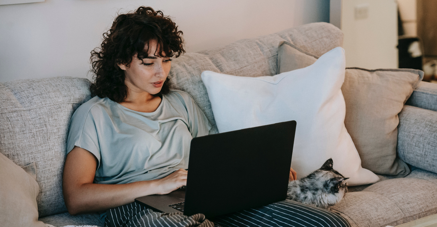 Eine Frau hat ihren Laptop auf dem Schoß und setzt mir einer Katze auf dem Sofa.