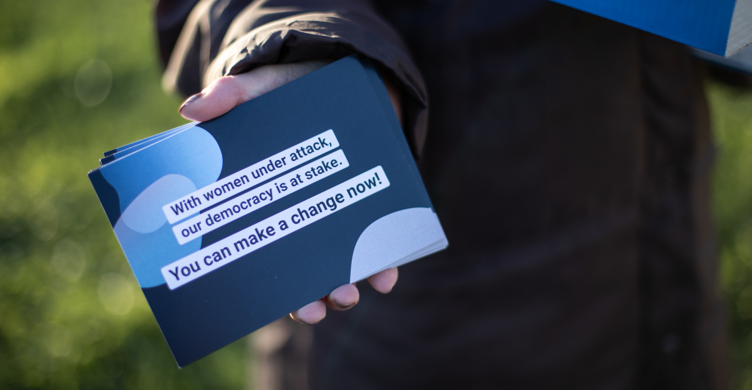 hateaid-was-du-tun-kannst-spendenaktion-starten Eine Frau, die eine blau-weiße Postkarte mit der Aufschrift: With Women under attack, our democracy is at stake. You can make change now.