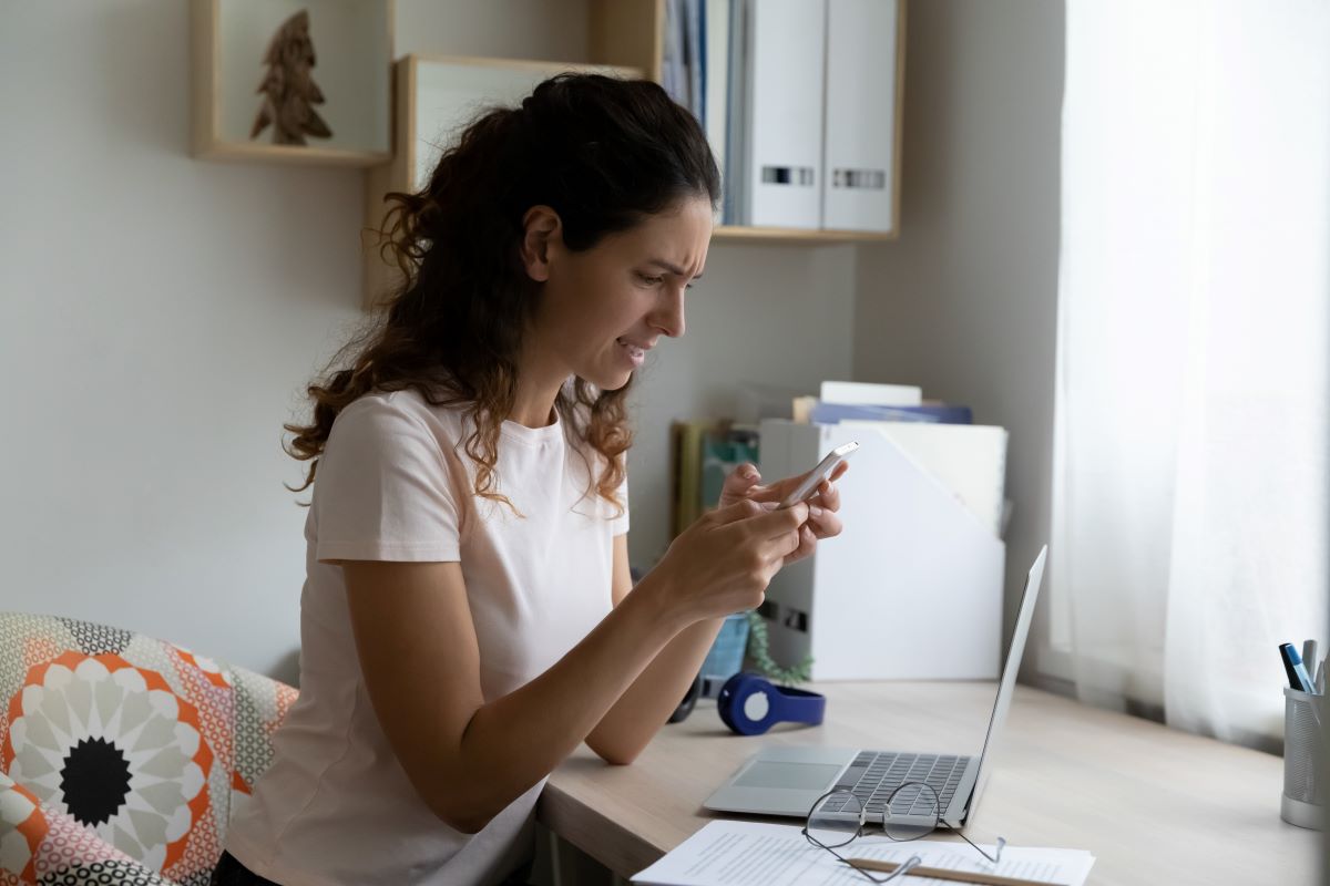 hateaid-gewaltfreie-kommunikation-artikelbild-2-c-shutterstock Person sitzt am Schreibtisch vor dem Laptop und guckt wütend auf das Handy.