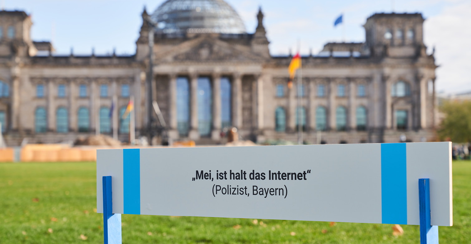 HateAid Hürdenlauf zum Gesetz gegen digitale Gewalt - Aktion vor dem Bundestag. Zu sehen ist im Hintergrund das Reichstagsgebäude sowie im Vordergrund ein Schild mit der Aufschrift "Mei, ist halt das Internet (Polizist, Bayern)"