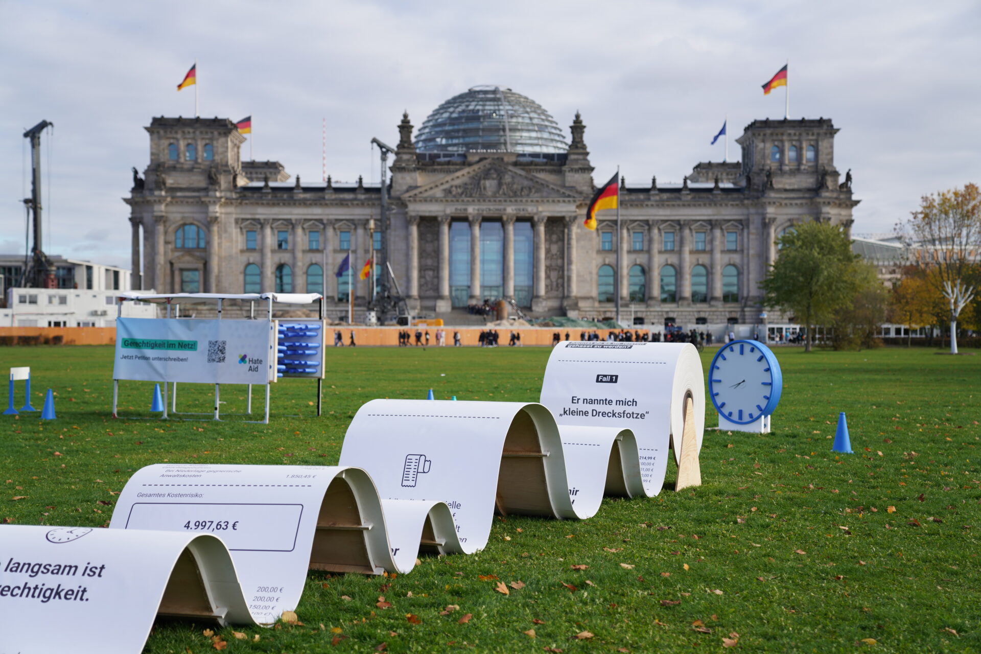 hateaid-huerdenlauf-und-rechnung-c-HateAid Hürdenlauf mit großer Rechnug, Uhr und einem Gerüst mit Banner zur Petition vor dem Bundestagsgebäude in Berlin.