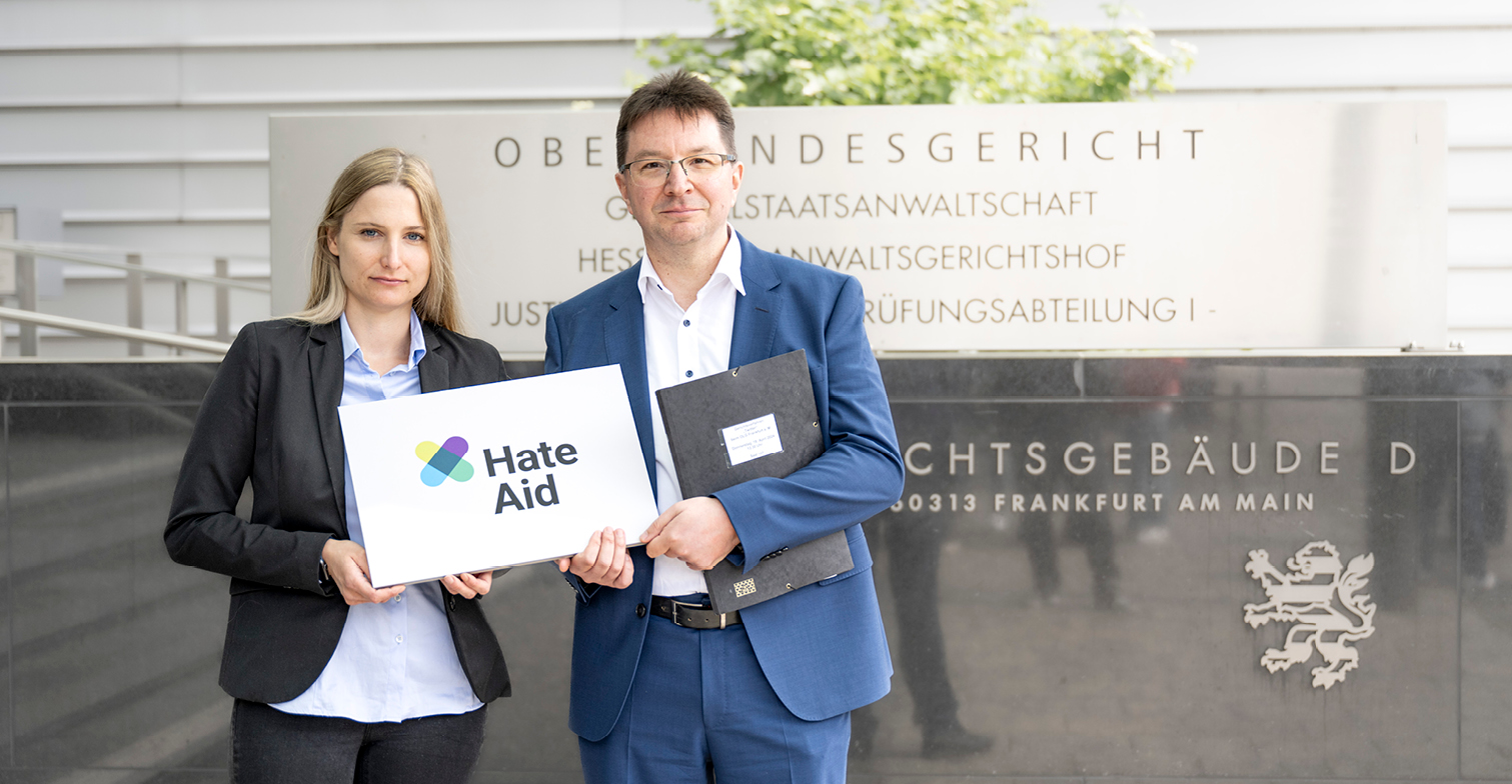 Josephine Ballon und Dr. Michael Blume vor dem Oberlandesgericht Frankfurt am Main. Beide halten gemeinsam ein weißes Schild mit der Aufschrift "HateAid" in den Händen.