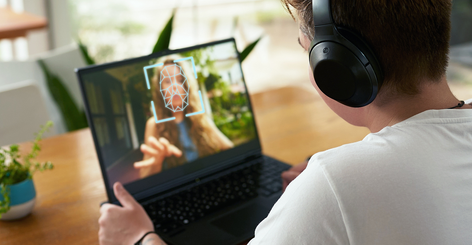 Deepfakes: Zu sehen ist eine Person, der über die Schulter geblickt wird. Die Person sitzt am Schreibtisch und hat einen Laptop vor sich, auf dem eine Frau mit einem Gesichtsscanner abgebildet ist.